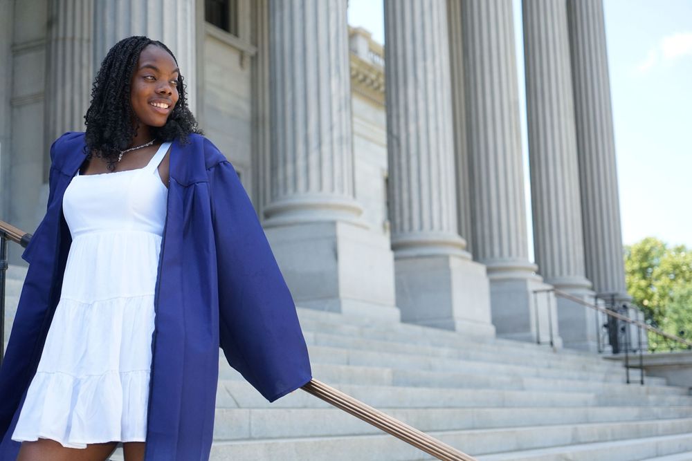 Young woman in white dress and blue graduation gown smiling on steps of a columned building.