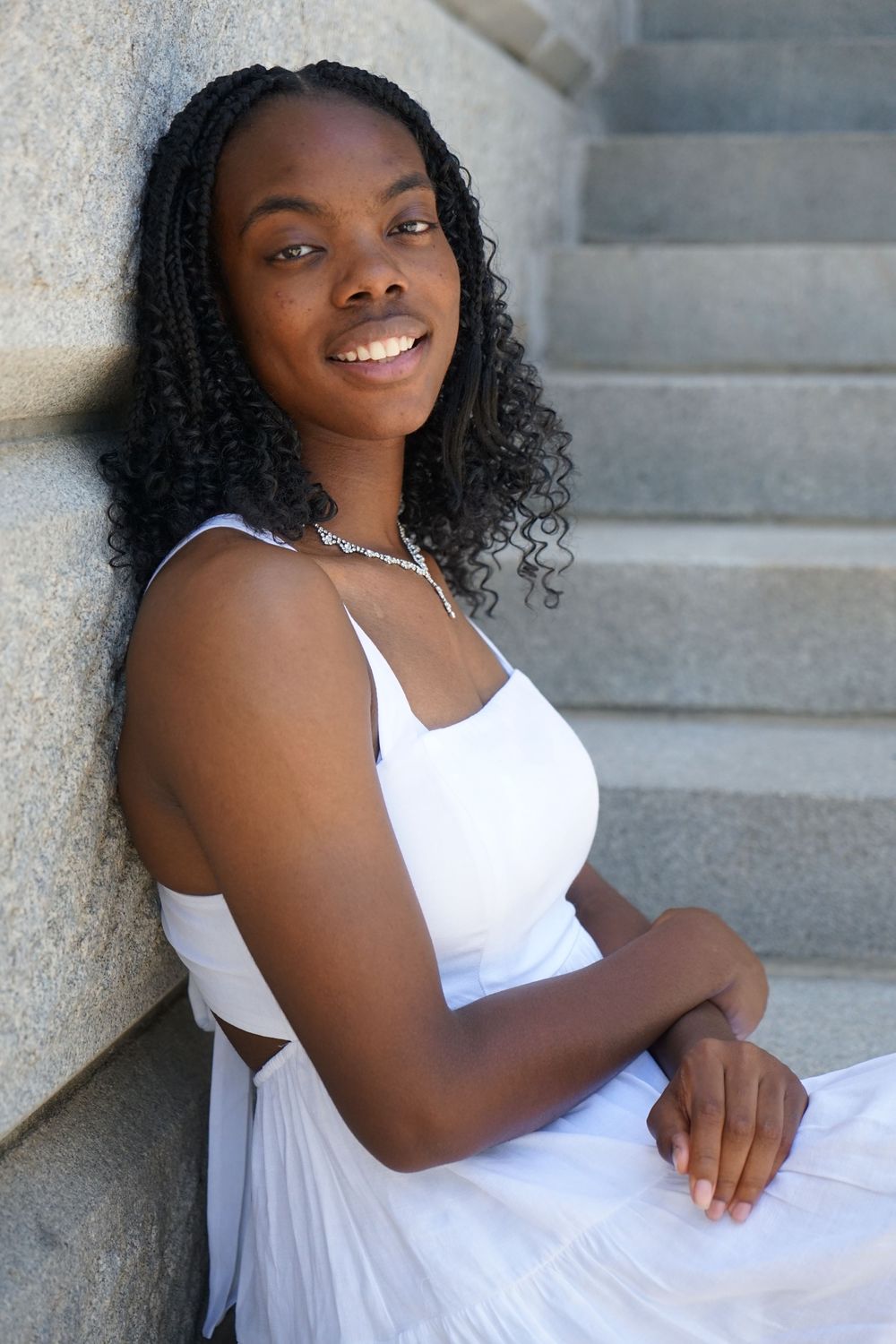 Young Black woman in white dress smiling while sitting on stone steps, leaning against a wall.