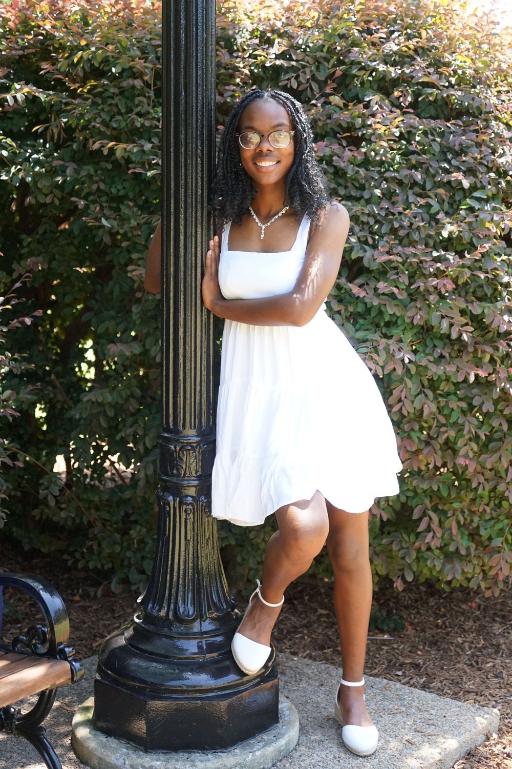 Young Black woman in white dress and glasses leaning against a lamp post, smiling outdoors near green and red foliage.