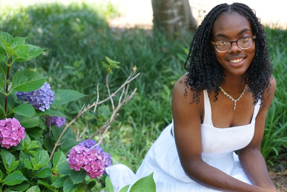 Young woman in white dress smiling beside pink and purple hydrangeas in a lush green garden.