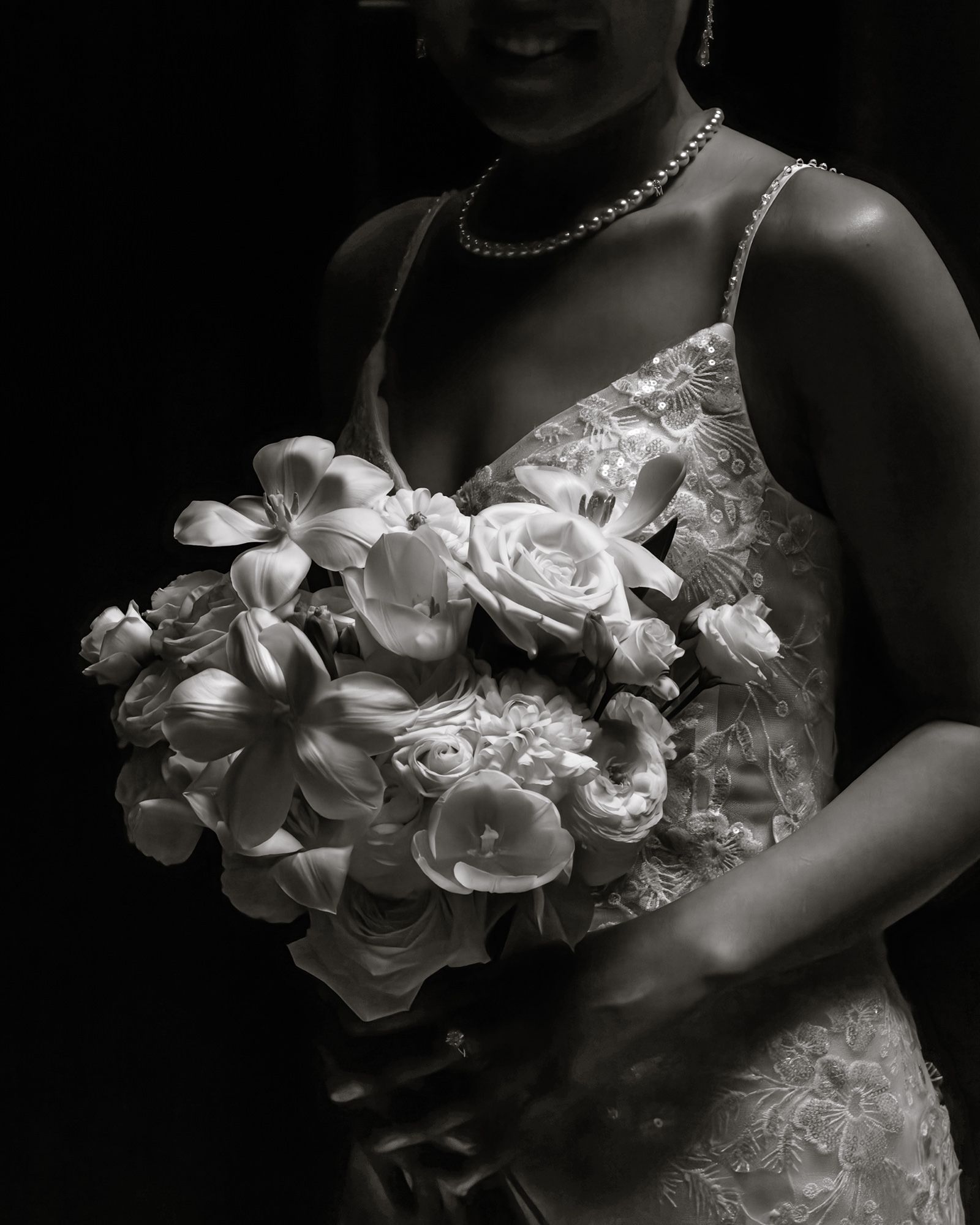 Black and white photo of a bride in lace gown holding a white floral bouquet with roses and lilies, wearing a pearl necklace.
