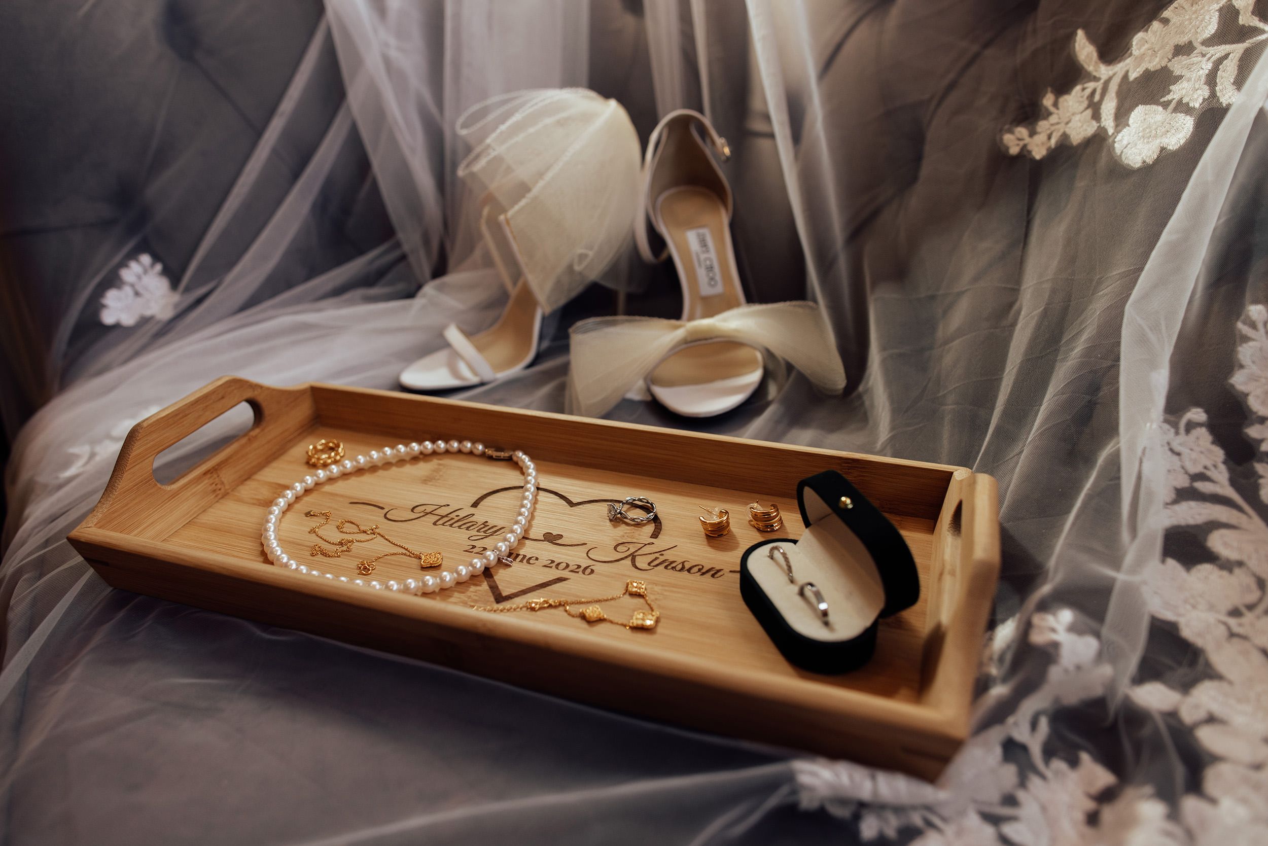 Elegant bridal flat lay with pearl necklace, earrings, heels, and engraved wooden tray on white lace veil.