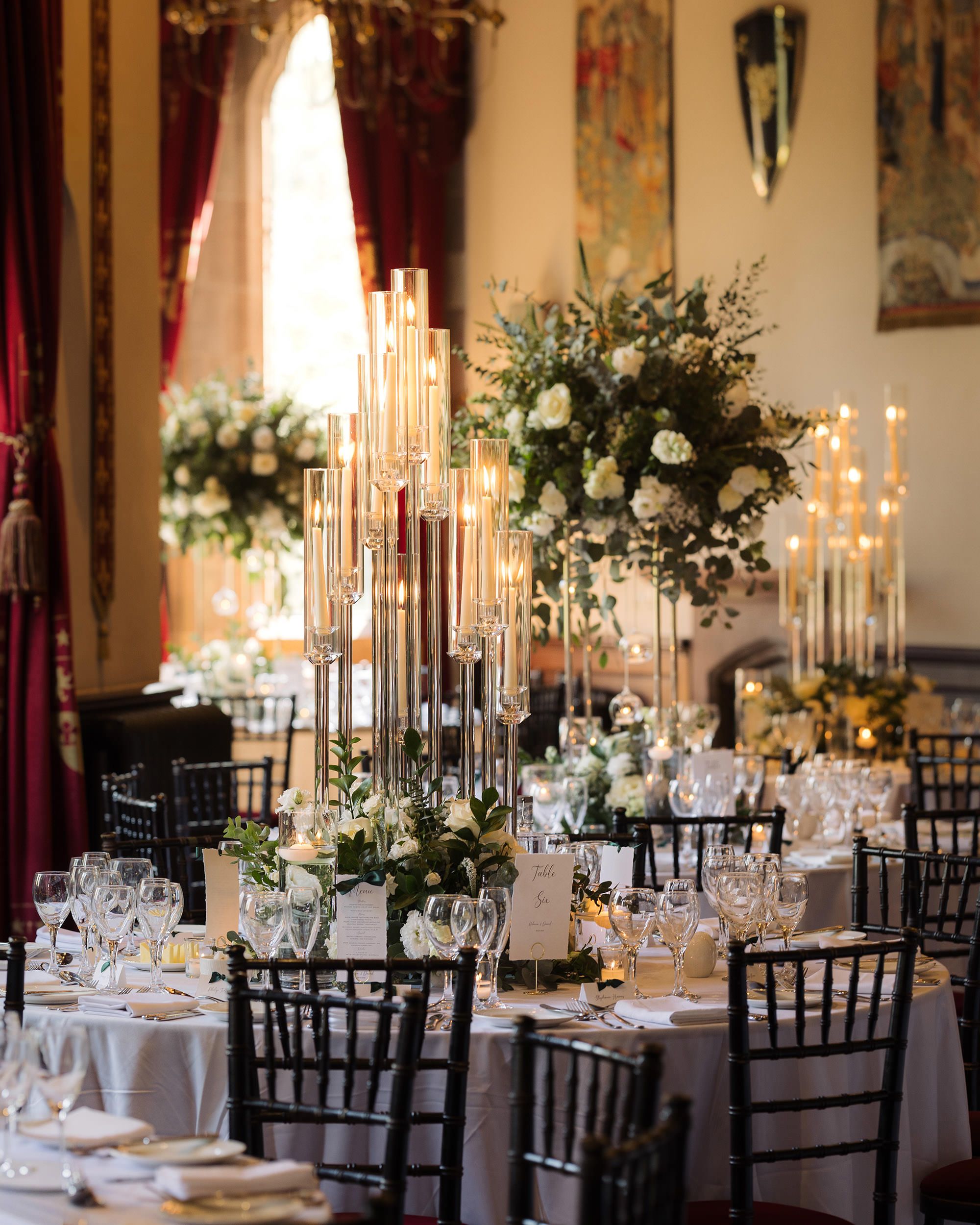Elegant wedding reception in a grand hall with tall candelabras, white floral centerpieces, black Chiavari chairs, and draped red curtains.
