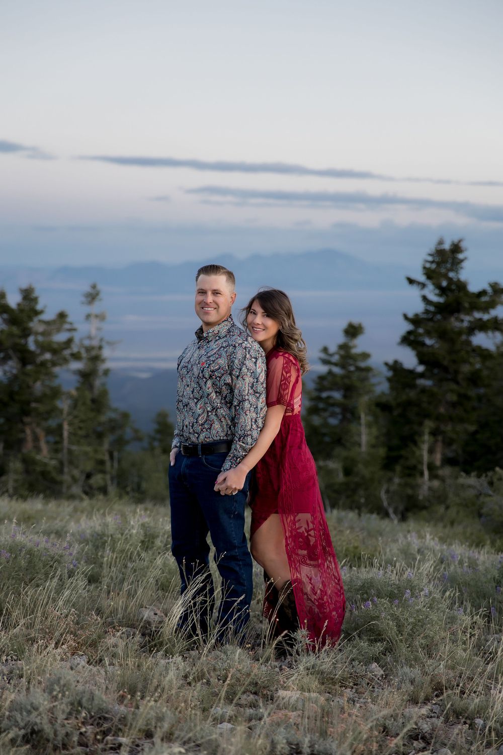 A couple in formal attire shares an intimate moment on a mountain overlook at dusk with scenic views in the background.