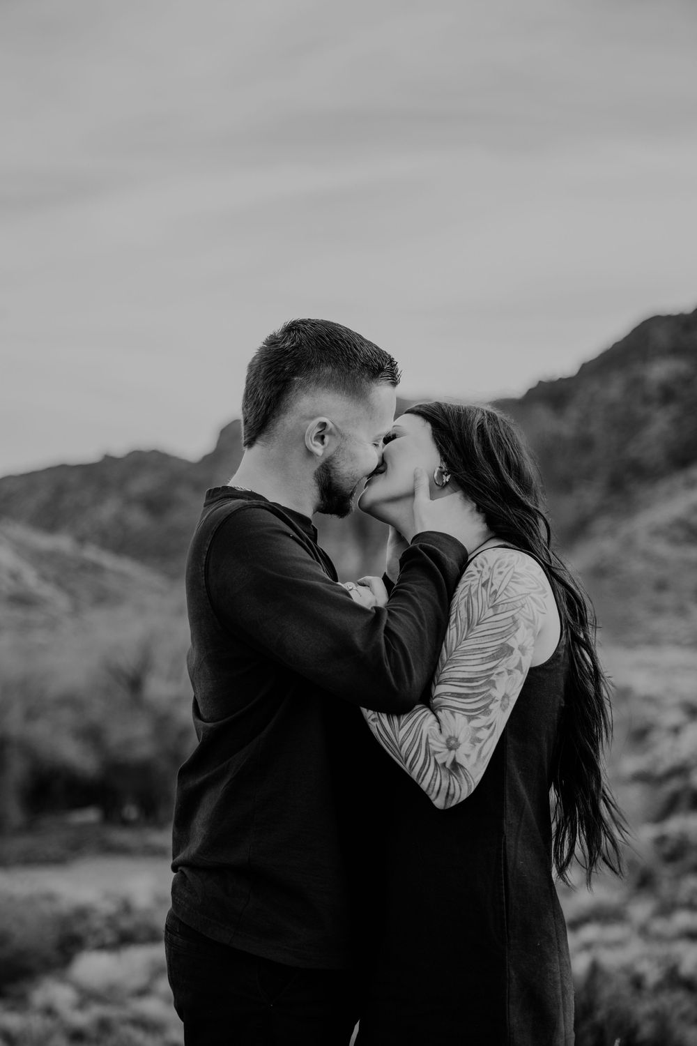 A romantic couple embraces and shares a tender kiss against a mountainous black and white landscape backdrop.