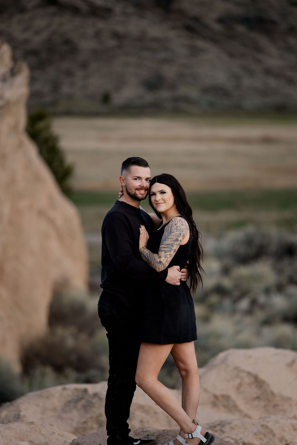 A romantic couple embraces on rocky terrain during a desert engagement photo session at sunset.