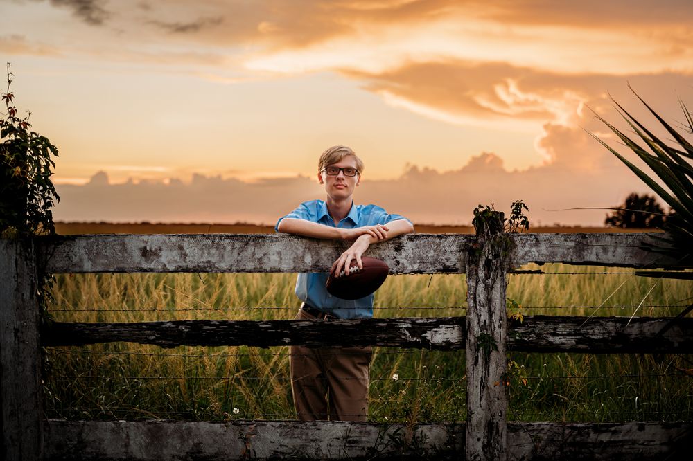 senior boy posing on a fence during sunset with a football