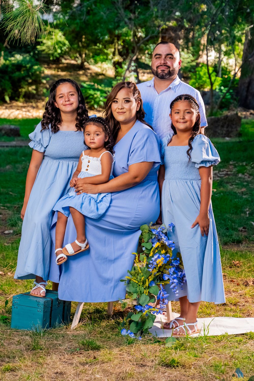 Family portrait in coordinating light blue outfits against lush greenery backdrop with blue flowers.