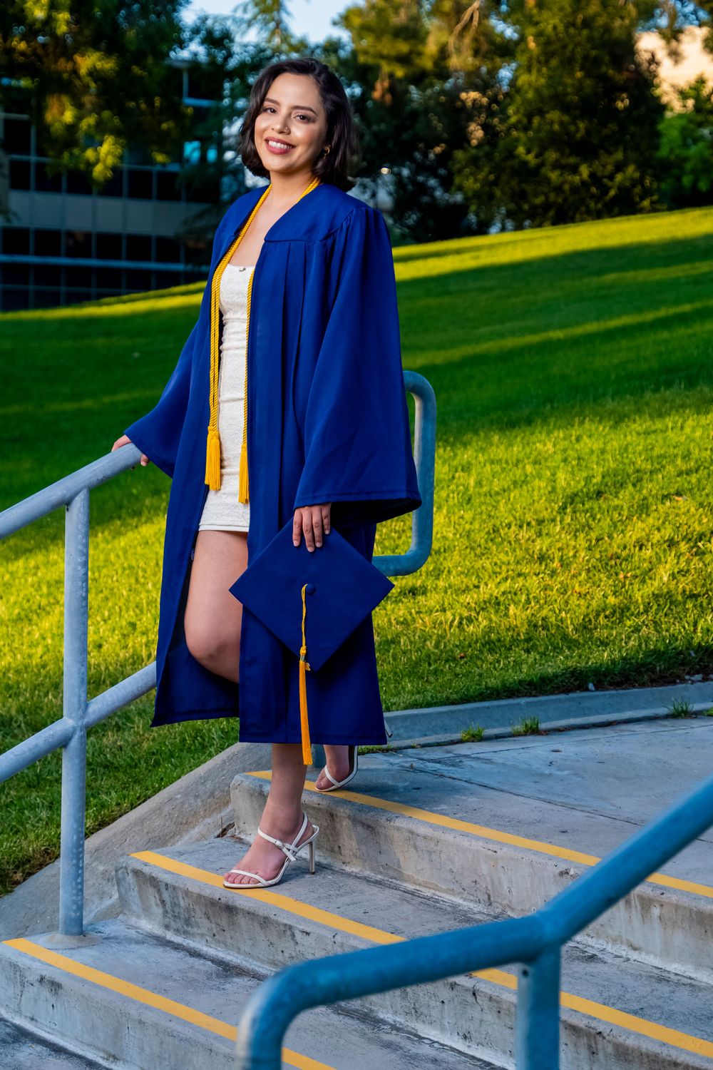 A graduate in a royal blue academic gown poses on concrete steps during golden hour.