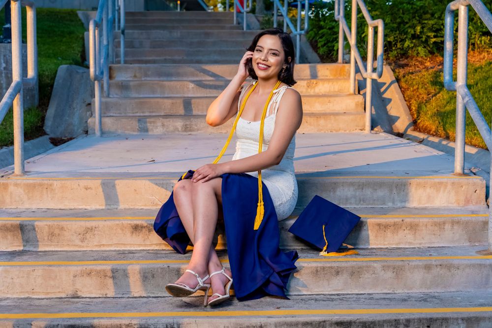 Graduate seated on concrete steps posing in blue graduation gown and white dress.
