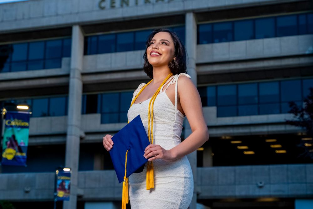 Graduate in white dress holding blue diploma folder outside Central Commons building.