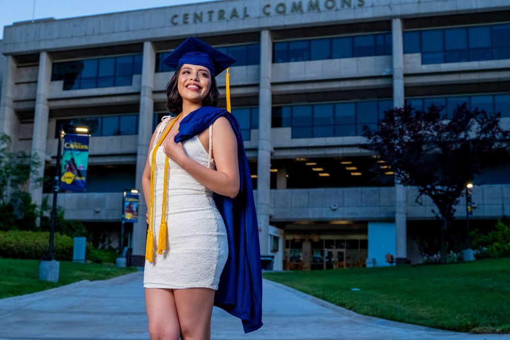 Graduate poses with diploma and graduation gown outside campus building at dusk.