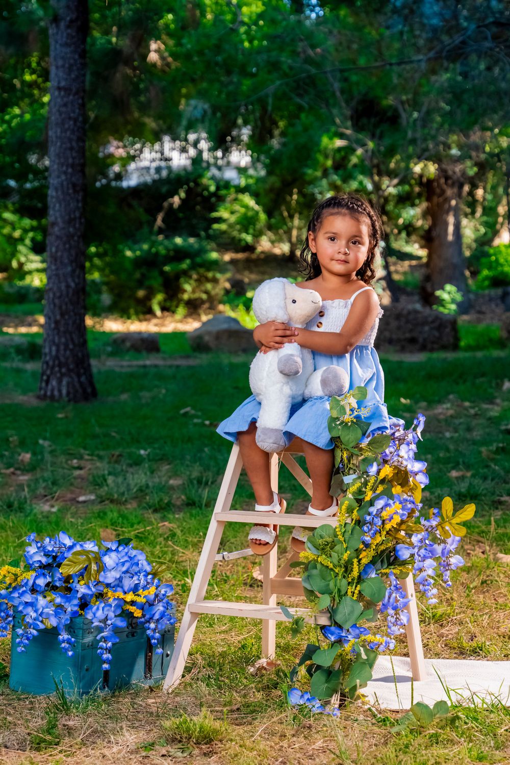 Young person in white dress holding teddy bear on wooden ladder with blue flowers.