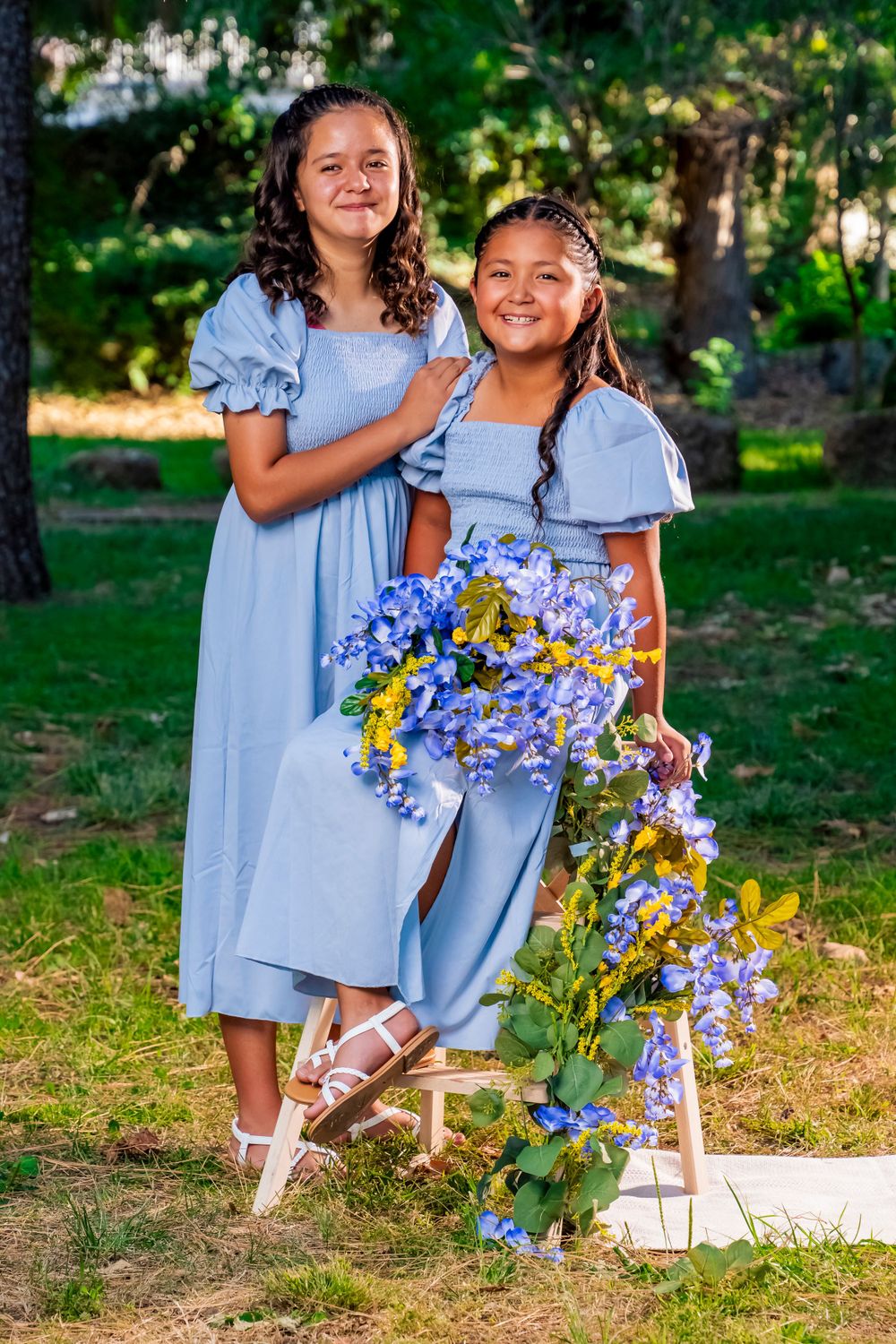Two people in light blue dresses pose with blue flowers in outdoor garden setting.