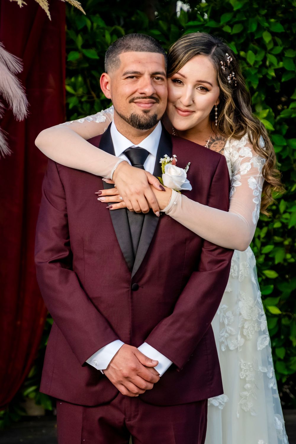 A wedding portrait featuring a couple in burgundy suit and lace gown against a green garden backdrop.