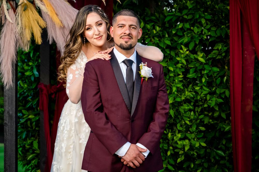 Newlyweds pose together at their wedding, with the groom in burgundy and bride in white lace.