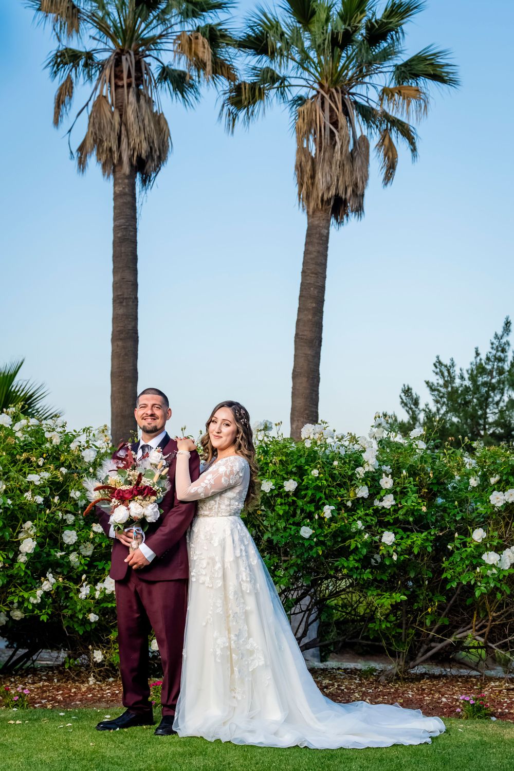 Wedding couple poses under tall palm trees with white rose bushes in a garden setting.