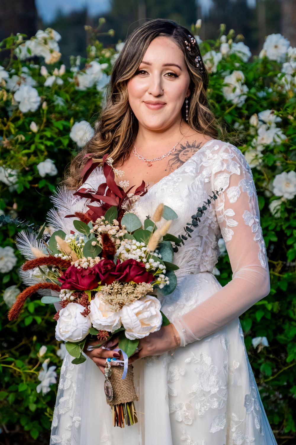 A bridal portrait showcasing a lace wedding gown and burgundy floral bouquet against white roses.