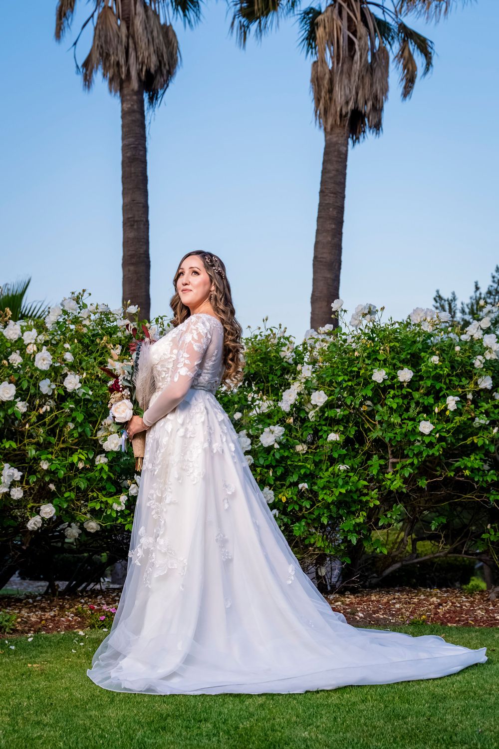 A bride in a white lace gown poses with her bouquet in a garden with palm trees.