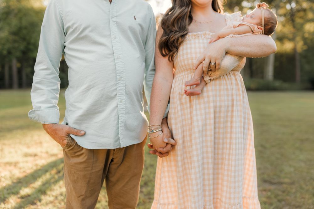 A couple stands together in a grassy field at sunset wearing light colored clothing and holding hands.