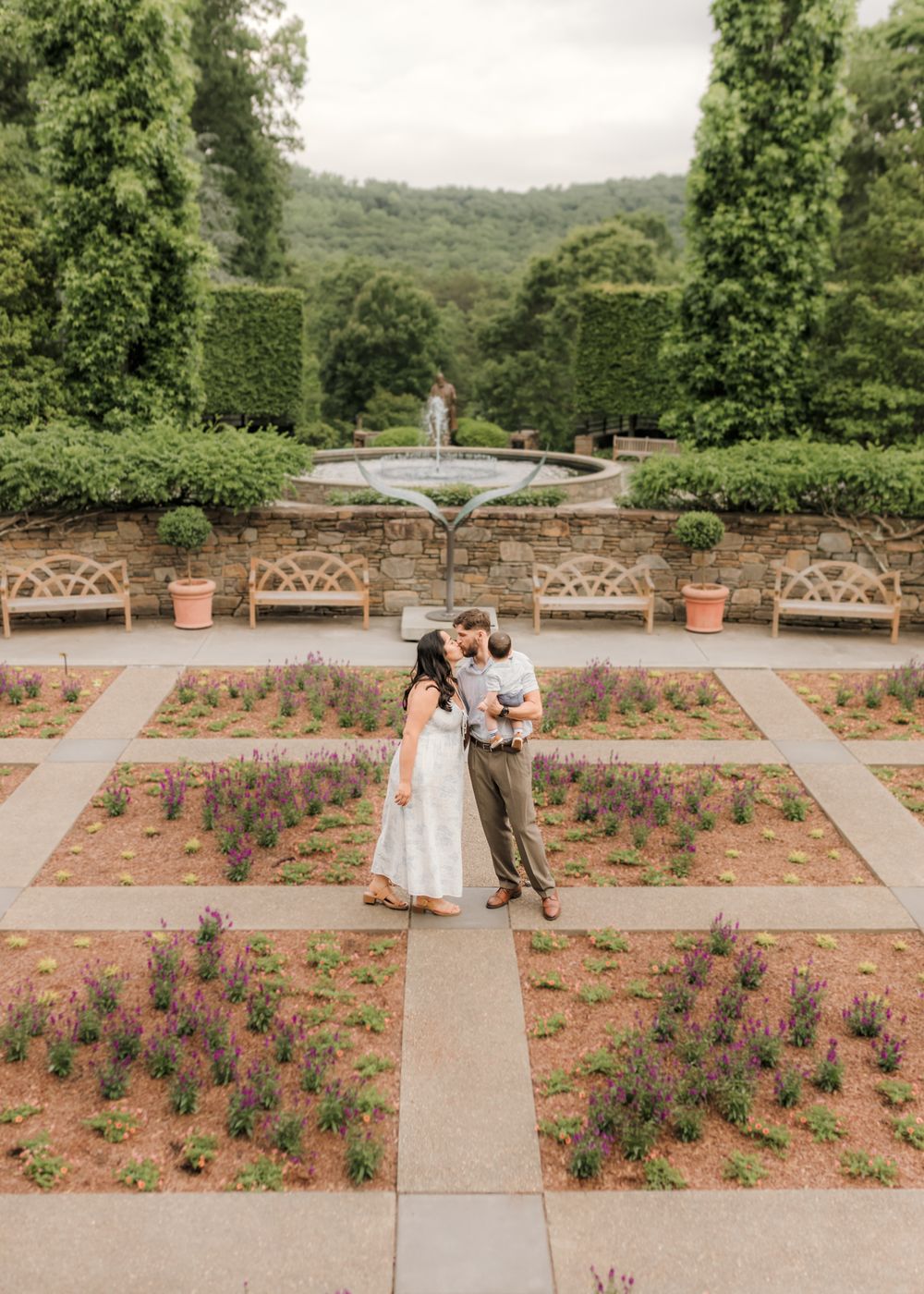 Couple embraces in a formal garden with geometric flower beds, stone fountain, and lush green hills in the background.