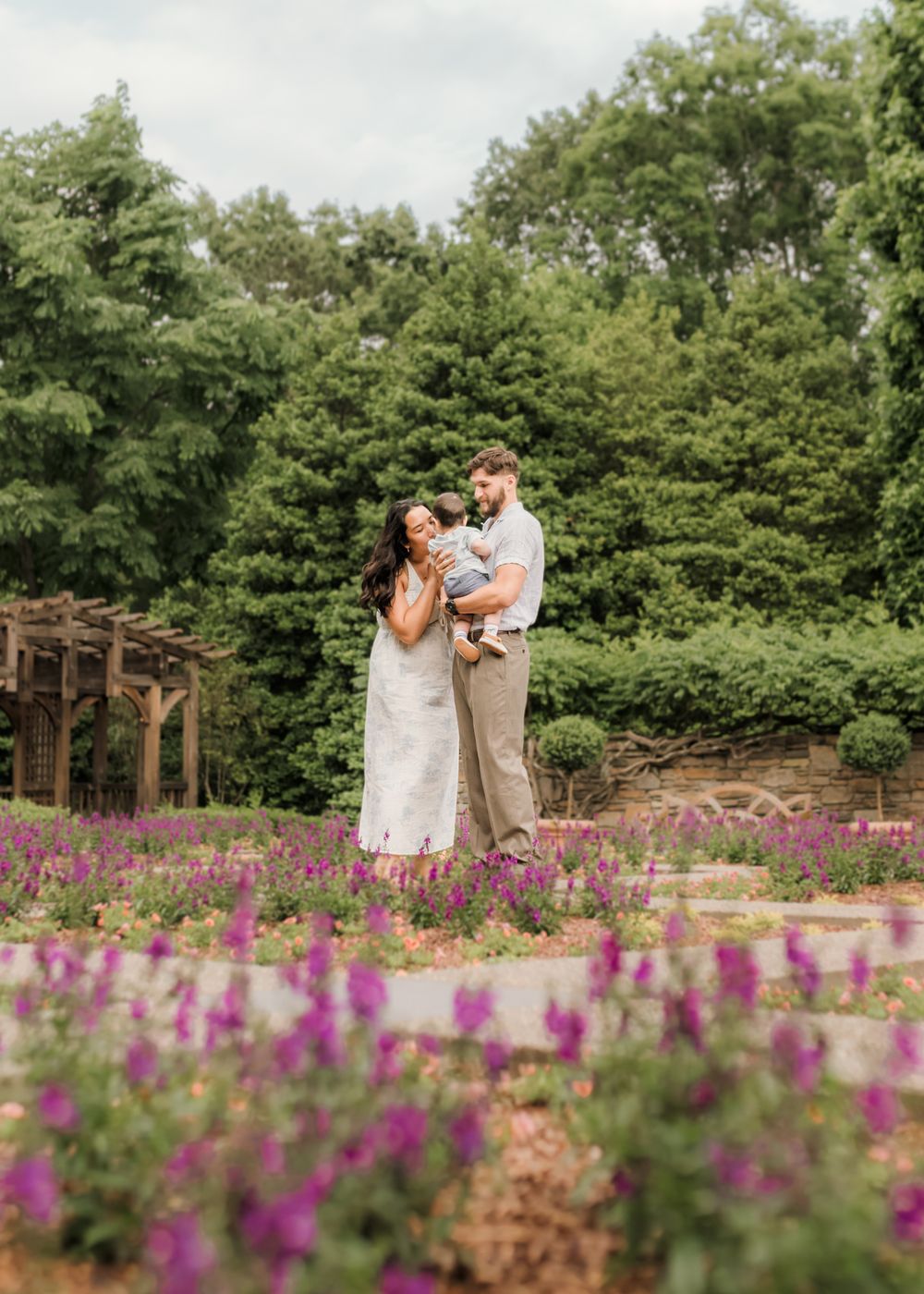 A couple holds their baby amid vibrant purple flowers in a lush garden setting.