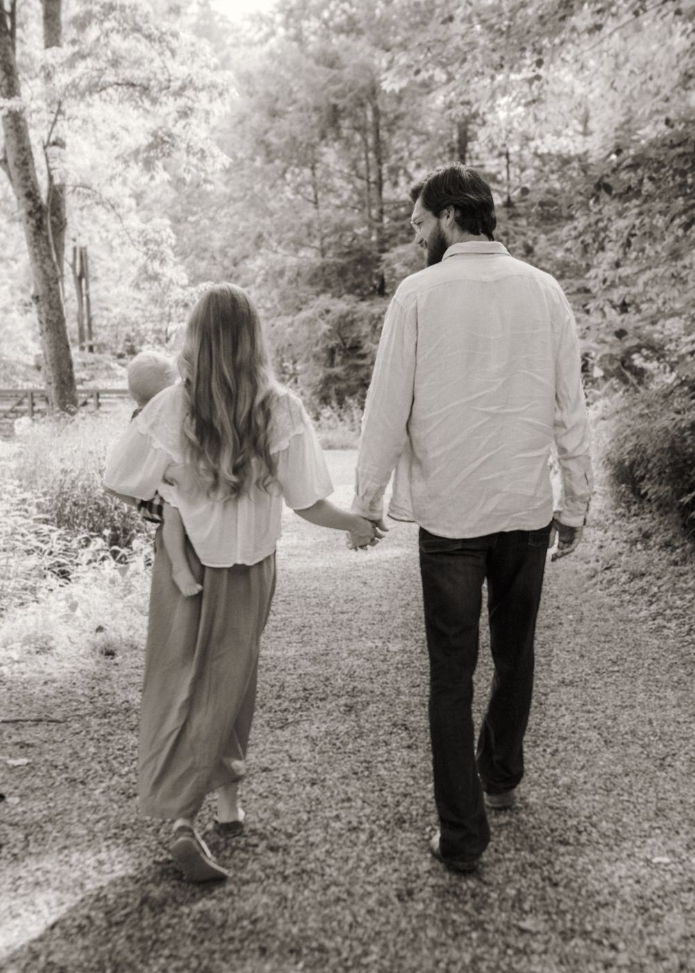 Black and white photo of a couple holding hands walking down a tree-lined path, woman in flowing dress.