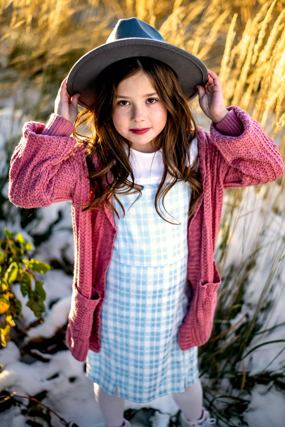 A little one in a gingham dress and pink cardigan poses while holding a wide-brimmed hat.