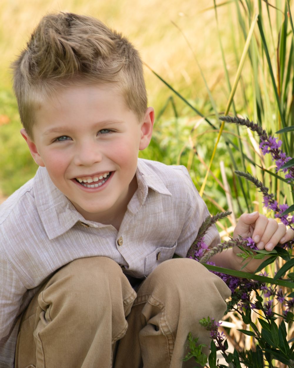 Young child sitting in grass with purple wildflowers on a sunny day wearing khaki pants and light colored shirt.