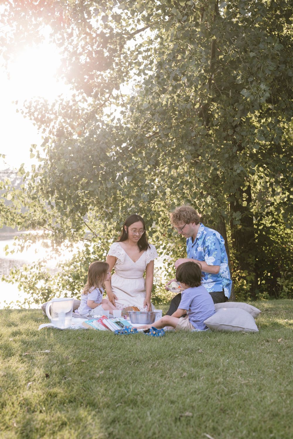 A family enjoys a sunny picnic together on a blanket in a grassy park with trees in the background.
