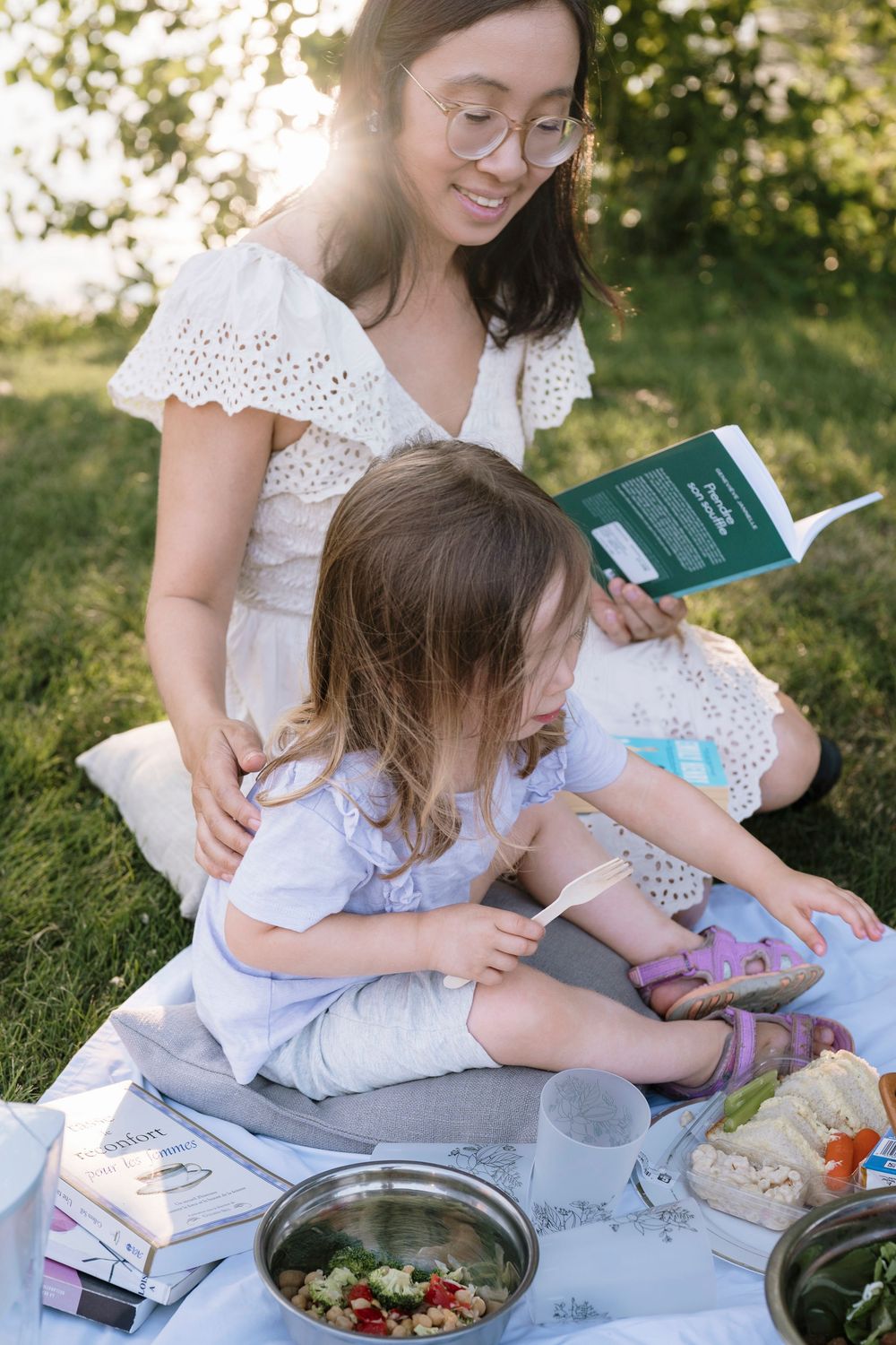 Parent and child sitting on picnic blanket outdoors reading books and enjoying fresh food together in summer.