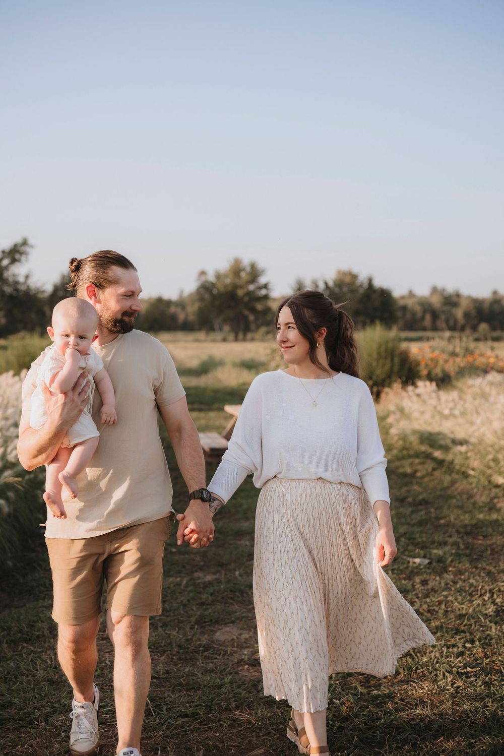 A couple and their baby walk together through a sunlit field wearing coordinated neutral-toned outfits.