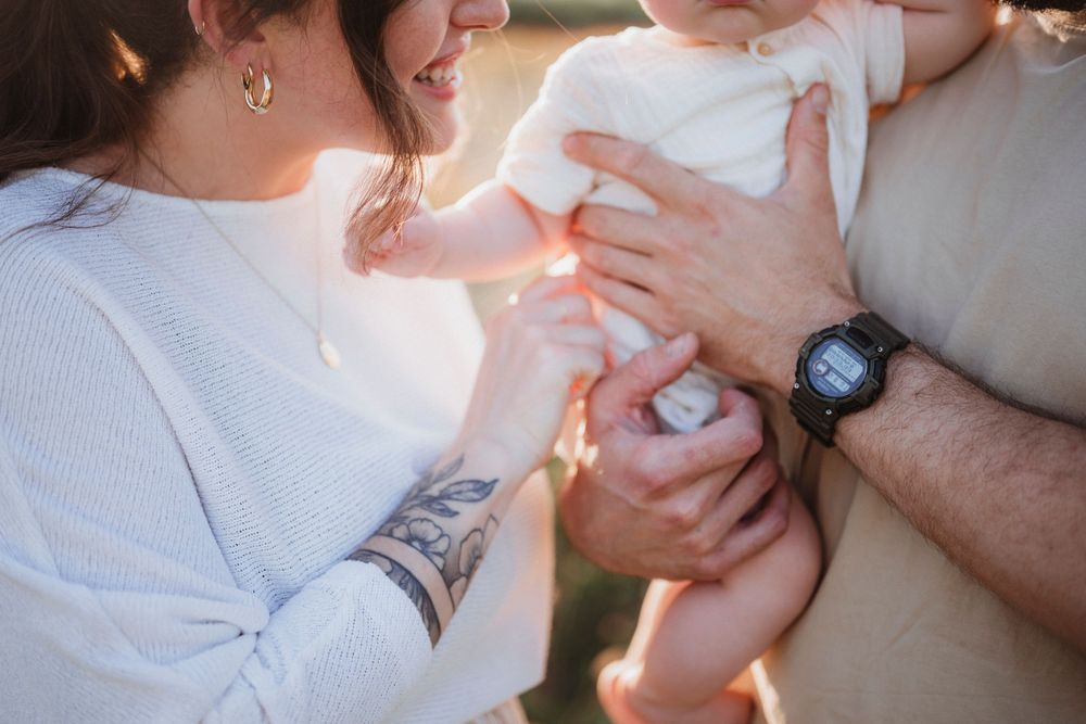 Hands tenderly hold a baby's feet in warm sunlight while wearing a white sweater and smart watch.