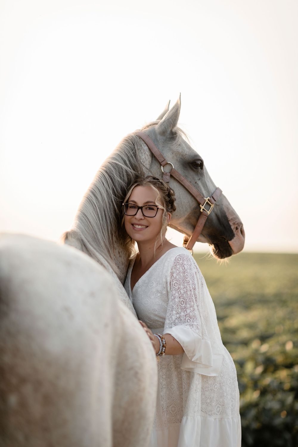 A white horse and someone in a lace dress share a tender moment in a sunlit field during golden hour.
