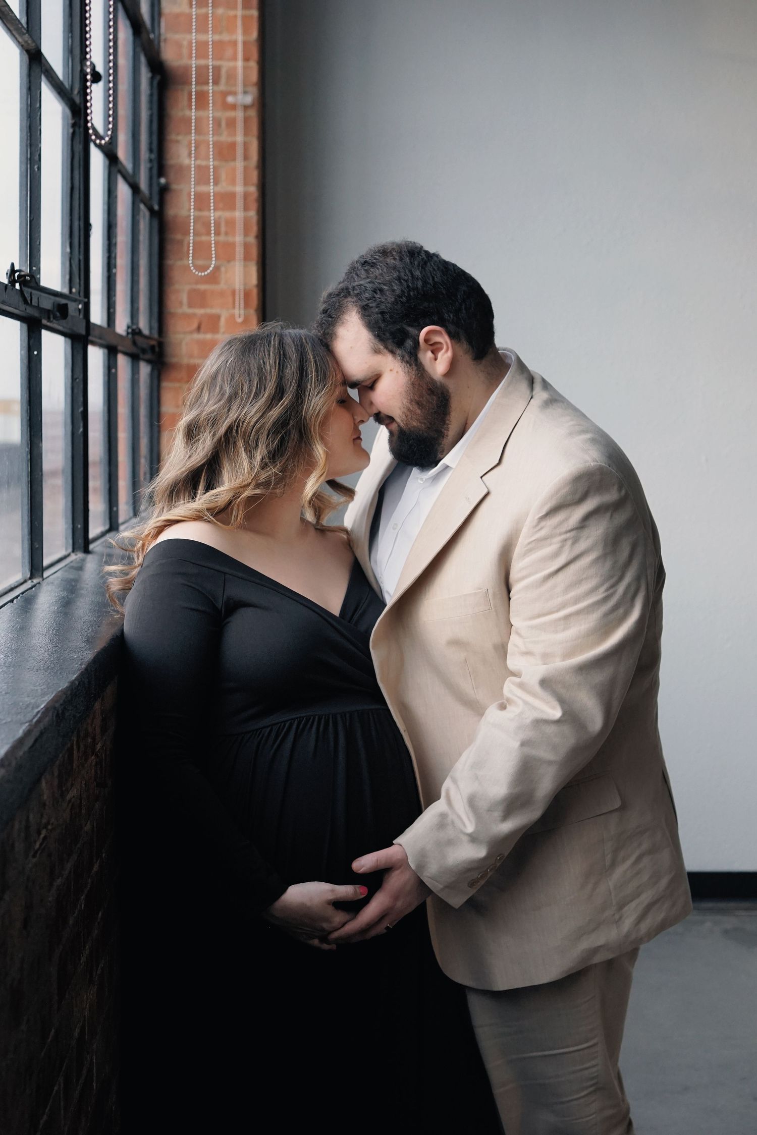 Elegant couple poses by industrial windows in formal attire for engagement photos.