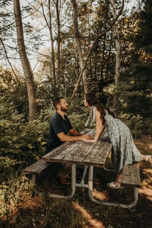 Two people sit at a rustic wooden picnic table in a sunlit forest setting having an intimate conversationMichigan Engagement Photographer |