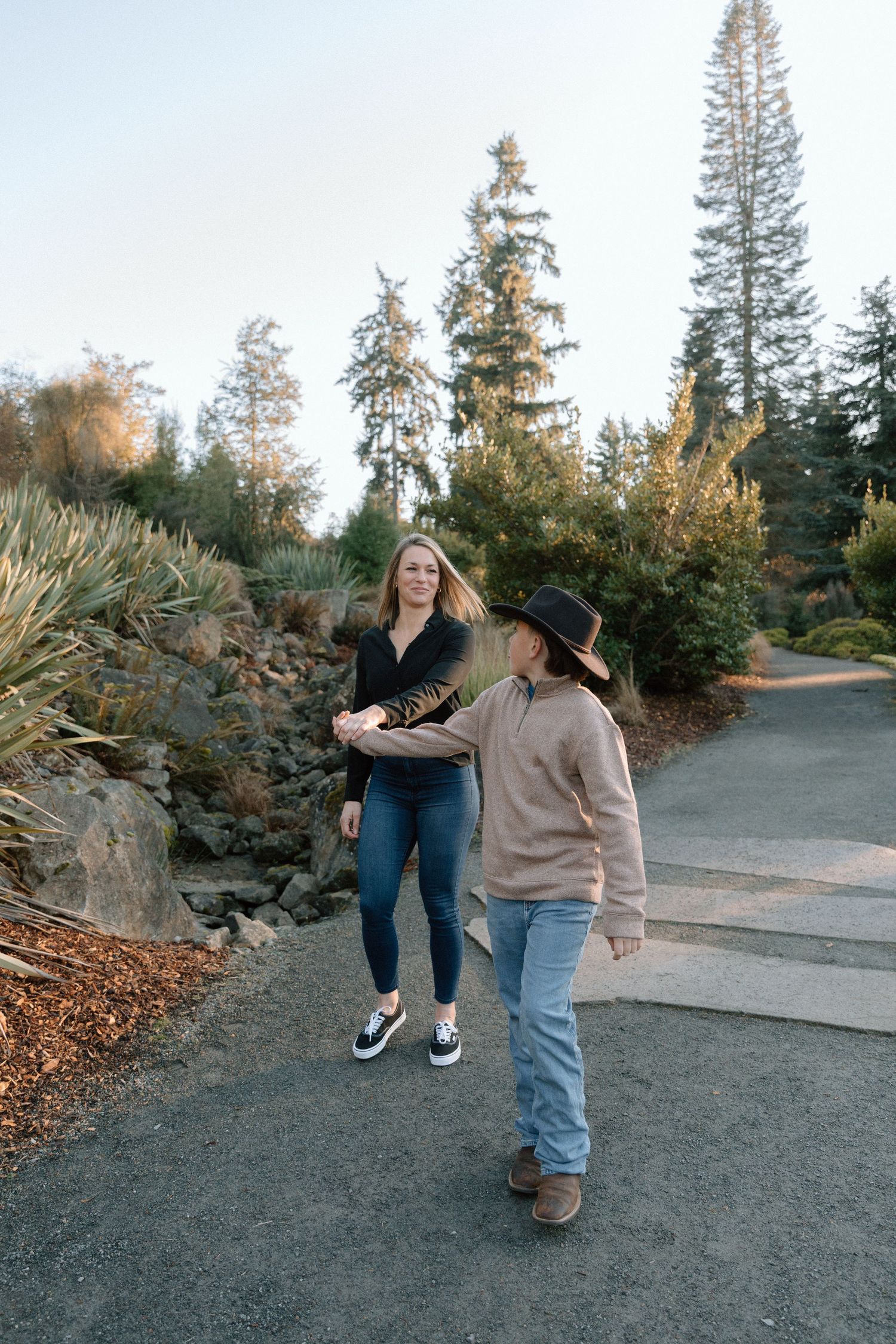Two women walking together on a garden path surrounded by evergreen trees and landscaped rocks.