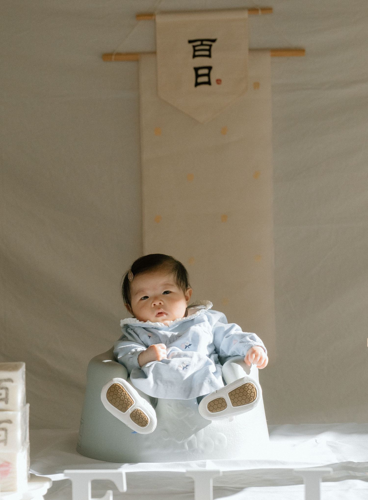 A baby in a light blue outfit sits in a small chair beneath a Japanese 100-day celebration banner.