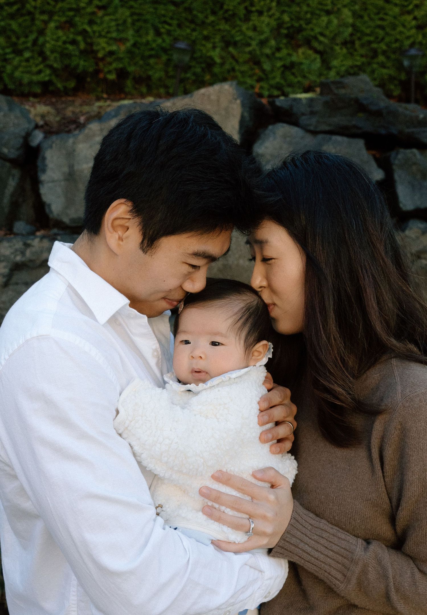 Asian couple tenderly holding their newborn baby, touching foreheads together in a loving family portrait outdoors.