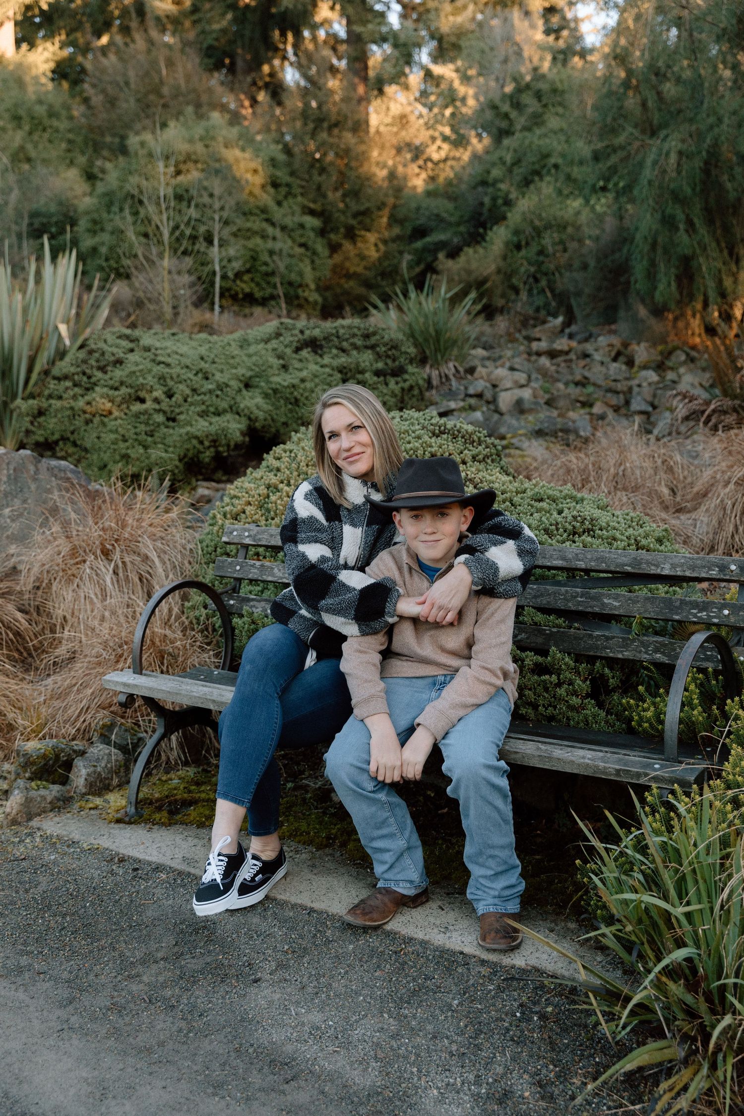 A woman and young boy in a hat sit together on a park bench surrounded by lush greenery.