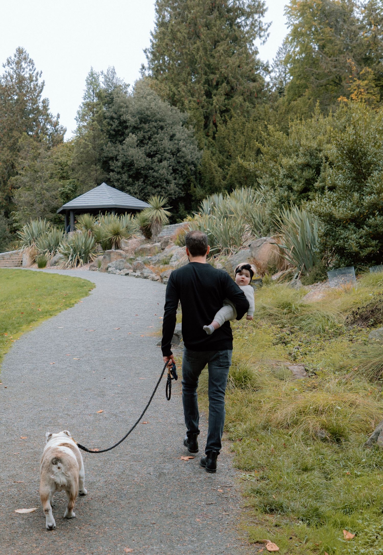 A dog and owner walking along a winding gravel path through a wooded park area on an autumn day.
