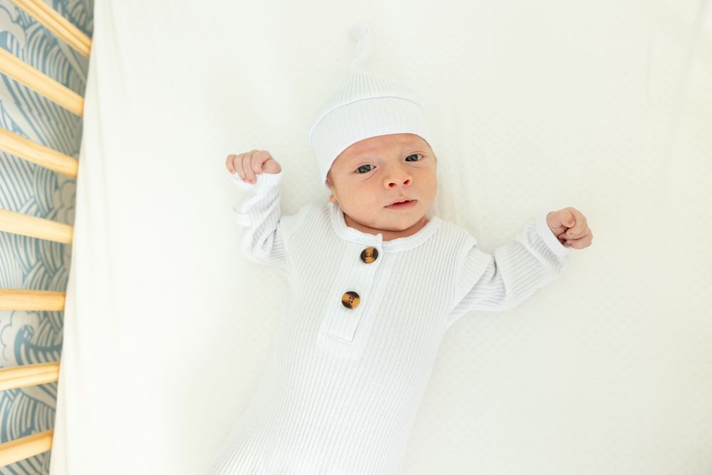 Baby wearing white outfit and cap lying on light colored background.