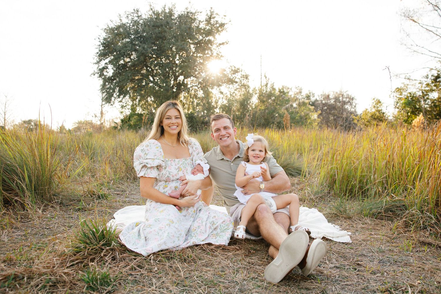 Family of four sitting outdoors at golden hour, parents with newborn and toddler daughter in white outfits, grassy field backdrop.