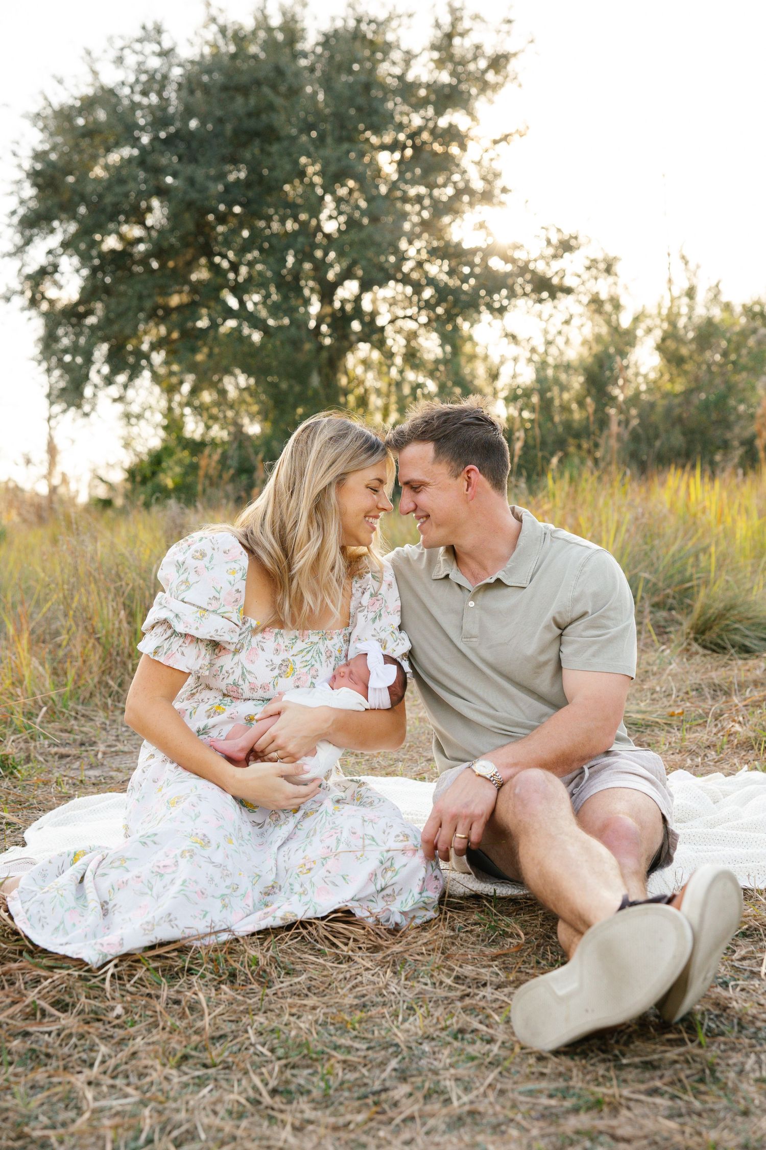 A couple sits on a blanket in a golden field, smiling at their newborn baby during an outdoor family photo session.
