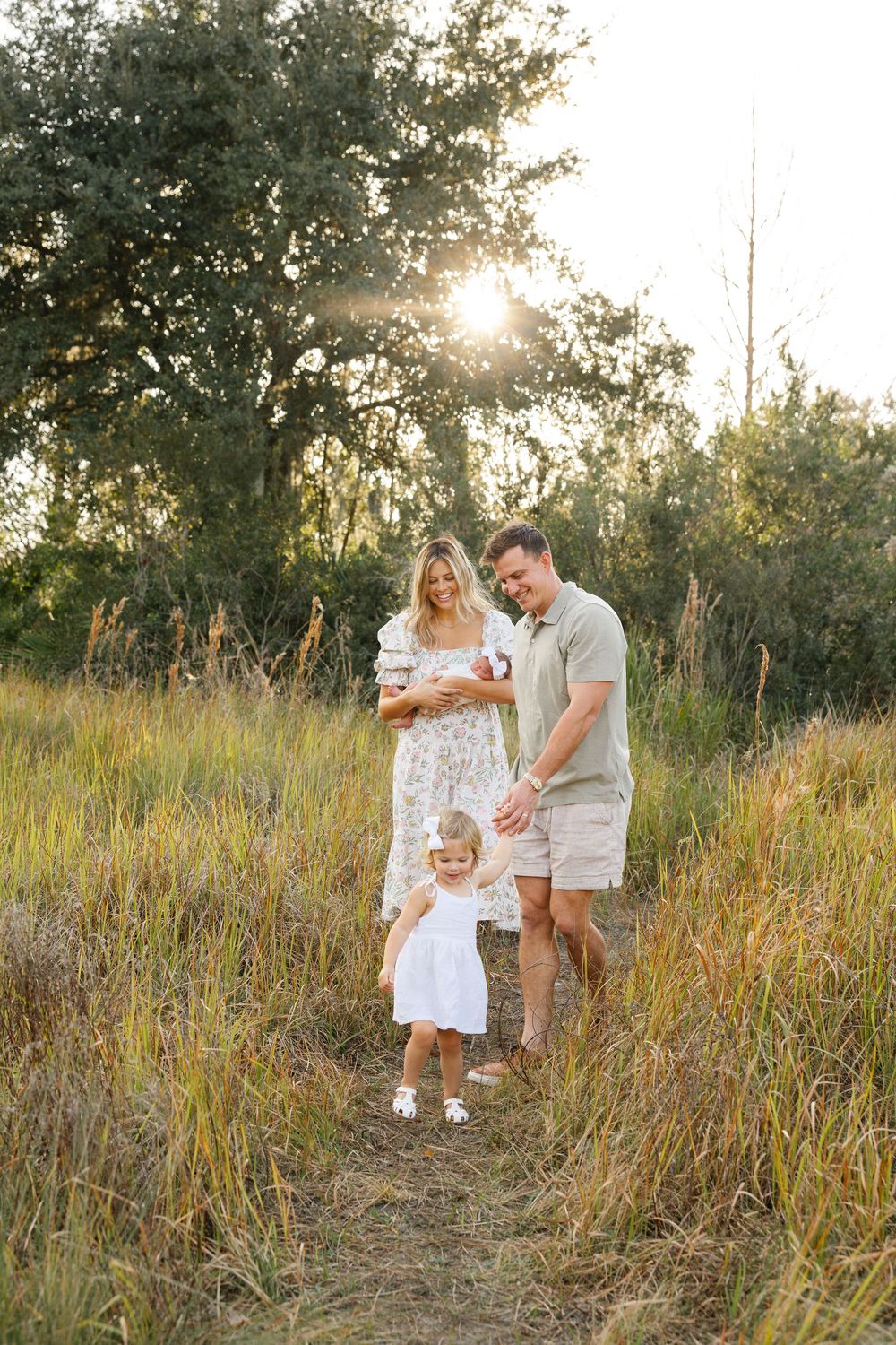Family of four walking in golden field at sunset, mother holding newborn, toddler girl in white dress leading the way.