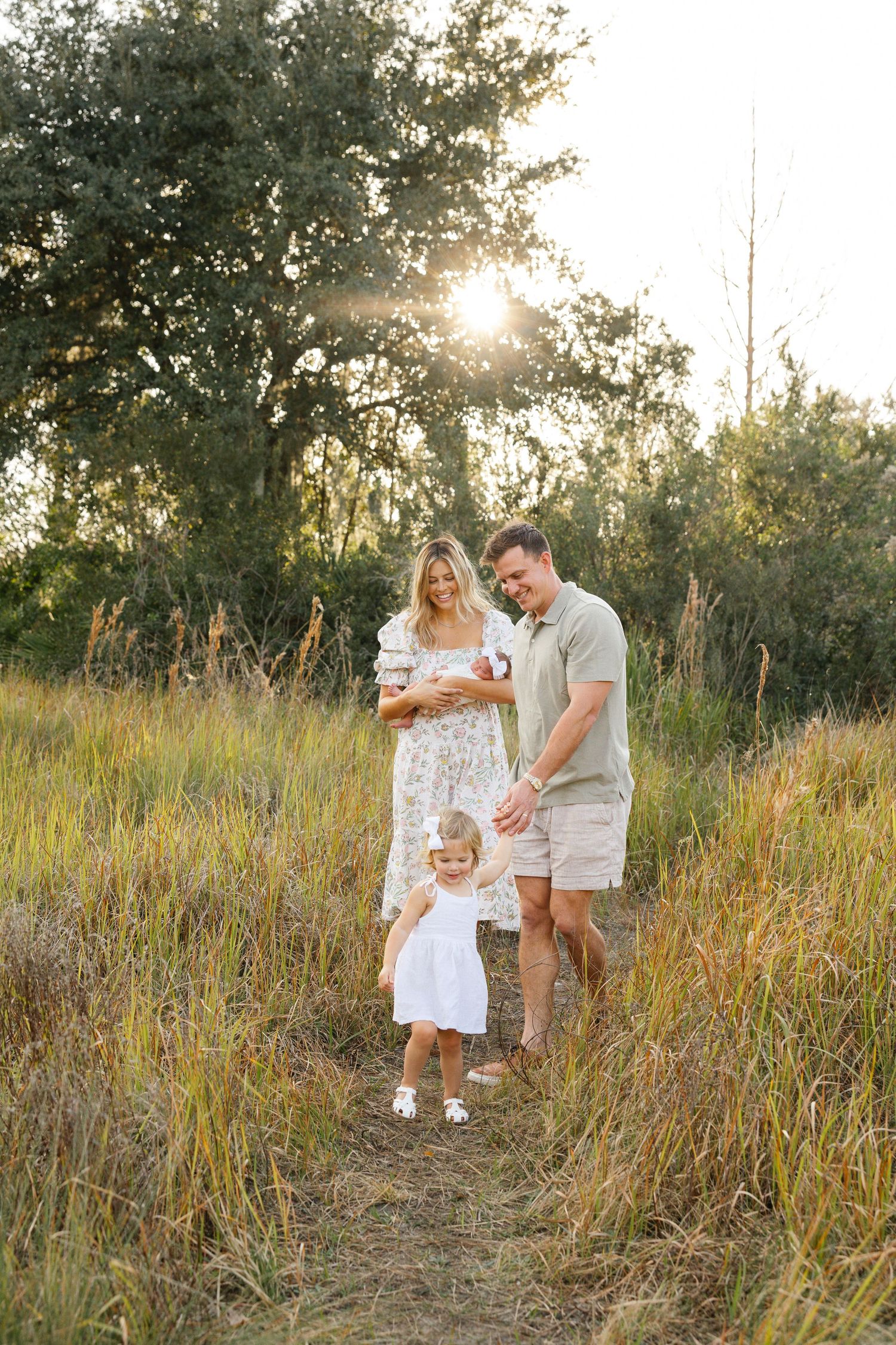 Family of four walking in golden field at sunset, mother holding newborn, toddler girl in white dress leading the way.