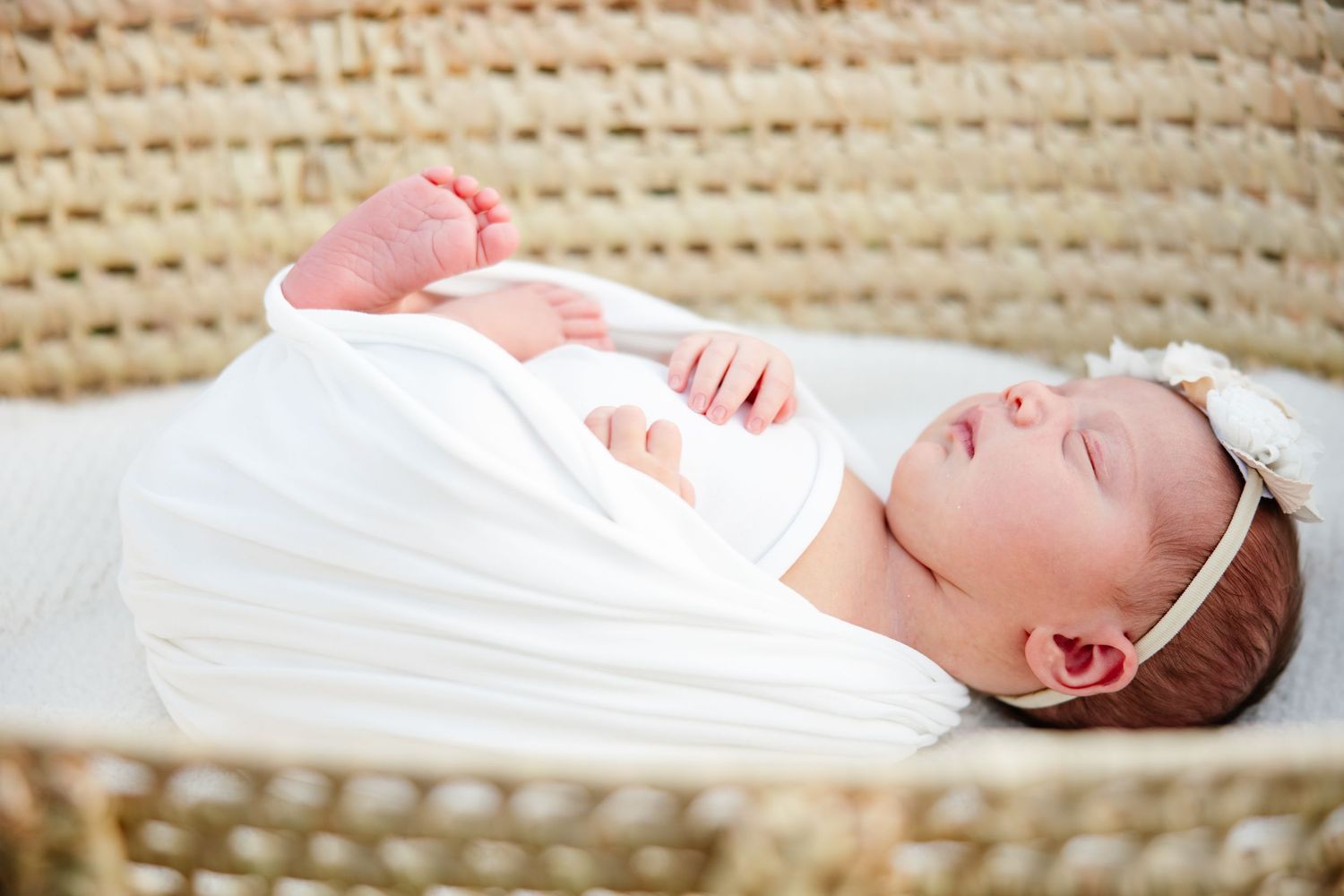 Peaceful newborn baby sleeping in a wicker bassinet, wrapped in white swaddle with a delicate floral headband.