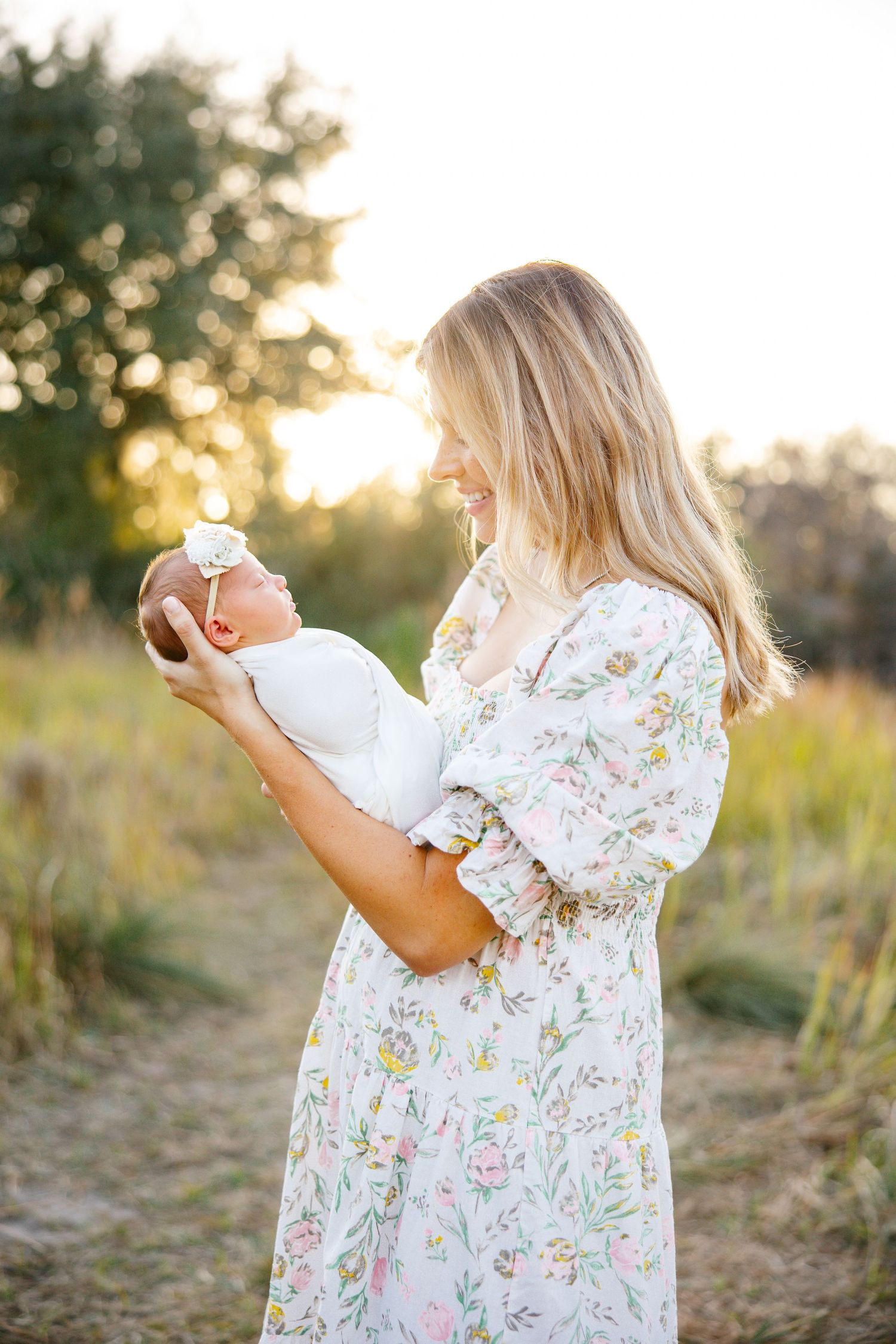 A blonde mother in a floral dress holds her newborn baby outdoors at golden hour in a sunlit field.