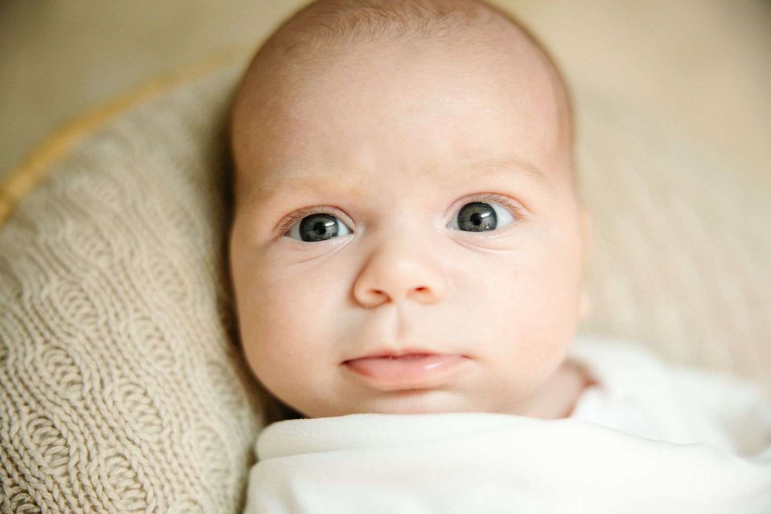 Close-up portrait of a newborn baby with blue eyes wrapped in white swaddle lying on knit blanket.