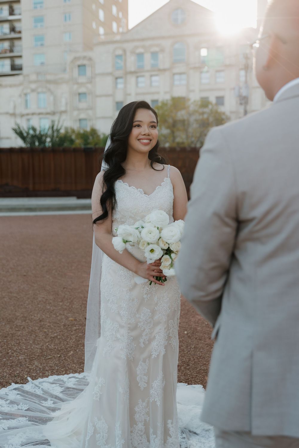 A bride in an elegant lace wedding gown holds a white bouquet during an outdoor ceremony at sunset.